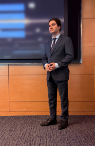 A person in formal attire stands near a podium in a conference room. Behind them is a large screen displaying a blurred presentation. The room has wood-paneled walls and carpeted flooring.