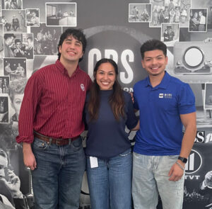 Three people are smiling in front of a wall decorated with vintage CBS-themed photographs and logos.