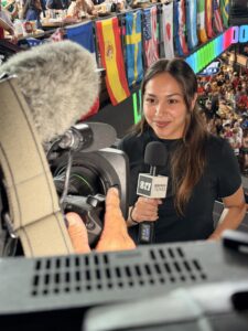 A person holding a microphone with a "Visit Texas" logo is speaking into a camera at a crowded event. Numerous international flags hang from the ceiling, including the Spanish flag.